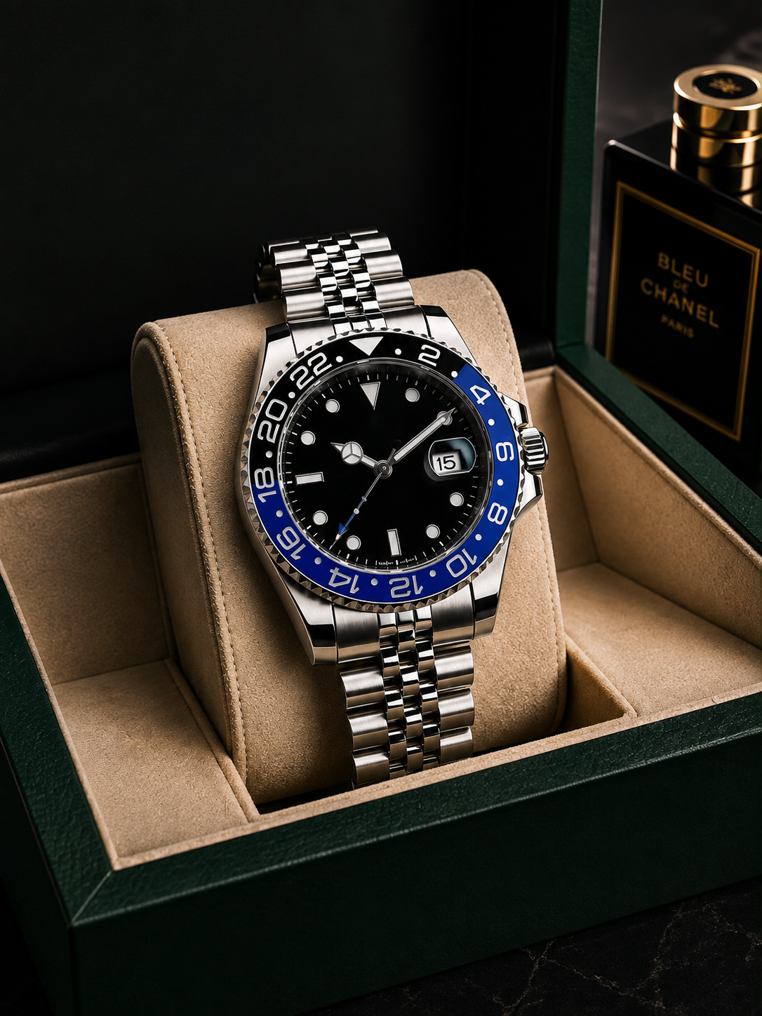 A stainless steel wristwatch with a black dial, blue and white two-tone bezel, and date window, displayed on a beige cushion inside a dark green watch box. A perfume bottle labeled Bleu Chanel Paris is partially visible in the background.