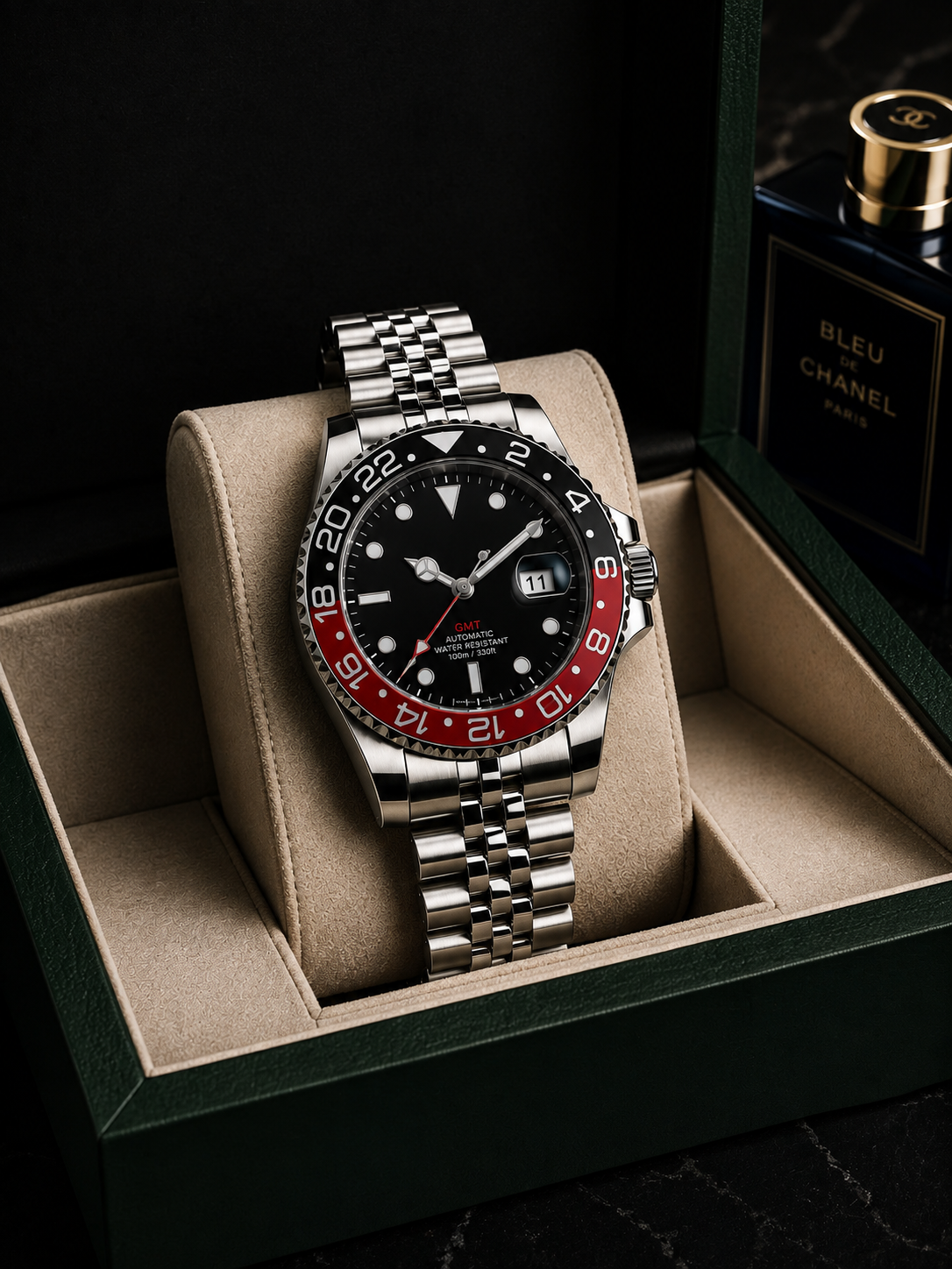 A stainless steel wristwatch with a black dial and red-and-black bezel sits displayed in a beige cushioned watch box. A dark green box and a perfume bottle labeled Bleu Chanel are partially visible in the background.