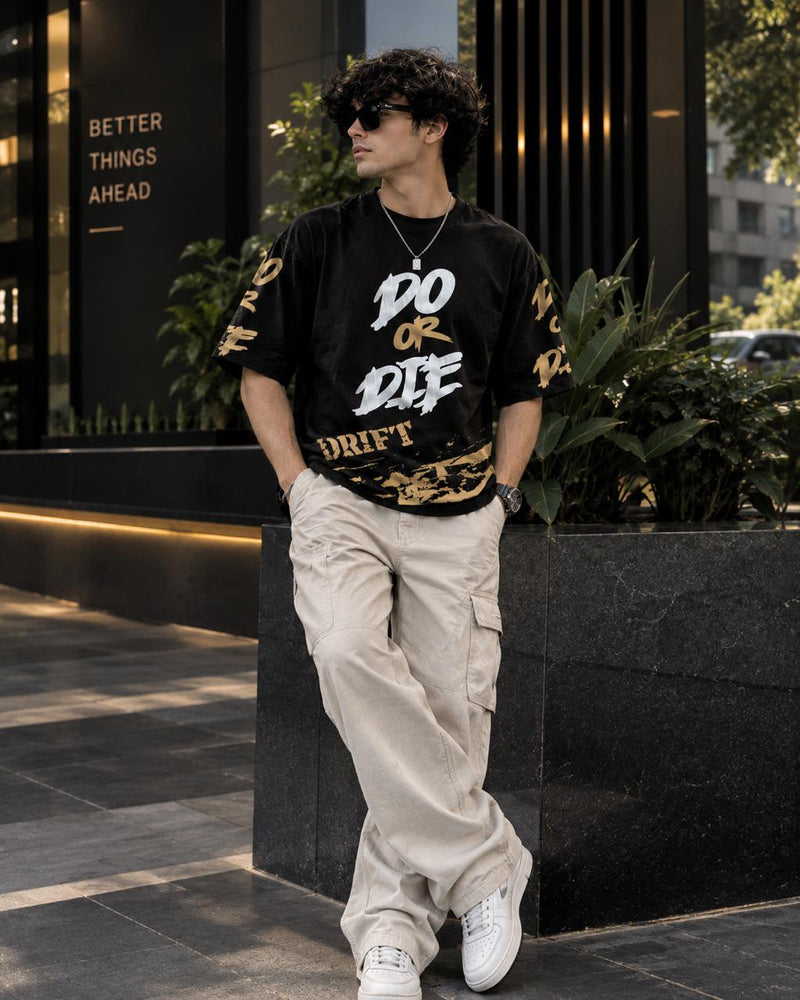 A man wearing a black graphic t-shirt with 'DO OR DIE DRIFT' in white and gold, paired with beige cargo pants and white sneakers, leaning against a modern black stone wall.