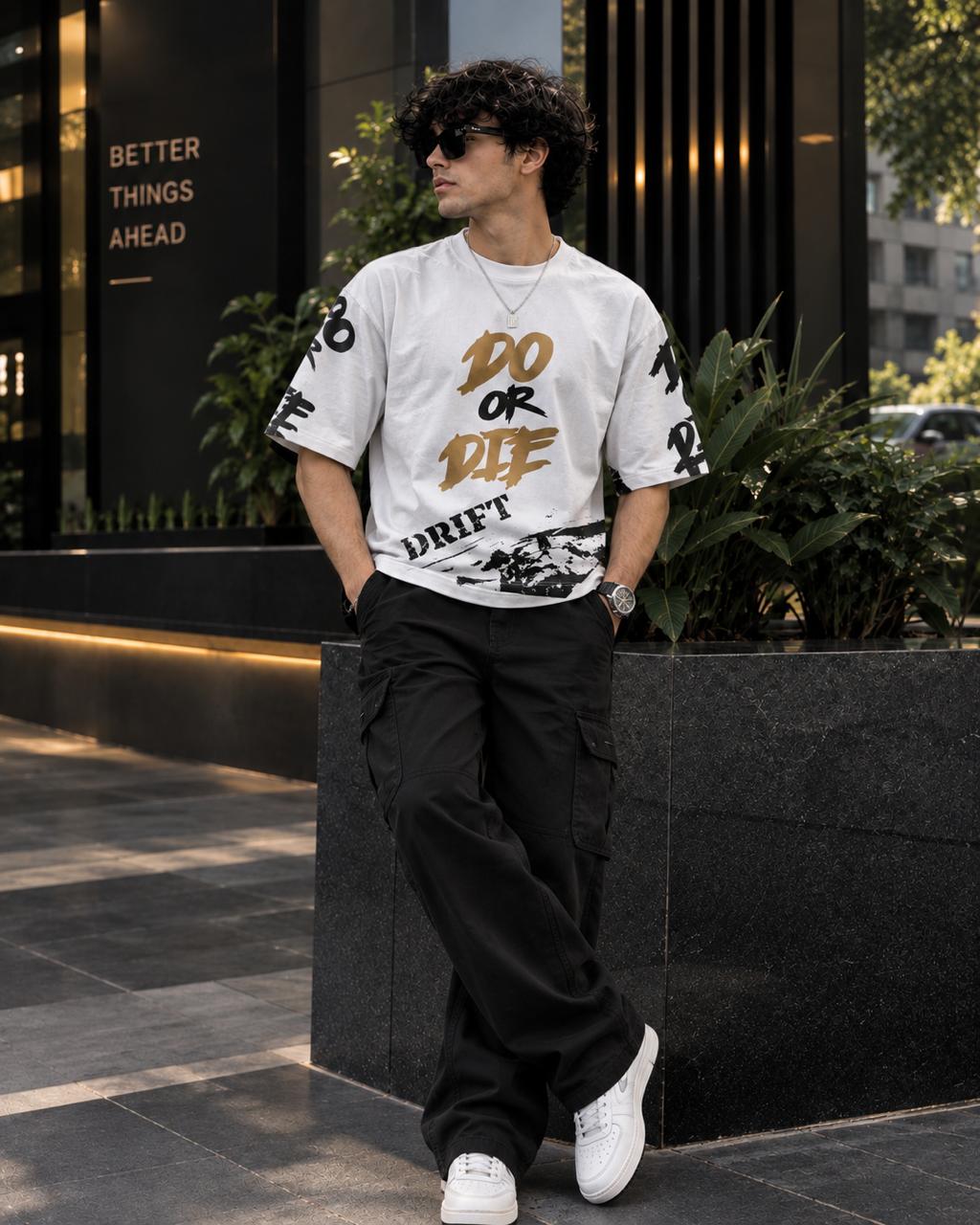 A man wearing a white graphic t-shirt with gold and black lettering that reads 'DO OR DIE DRIFT', paired with black cargo pants and white sneakers, standing outdoors in front of a modern building.