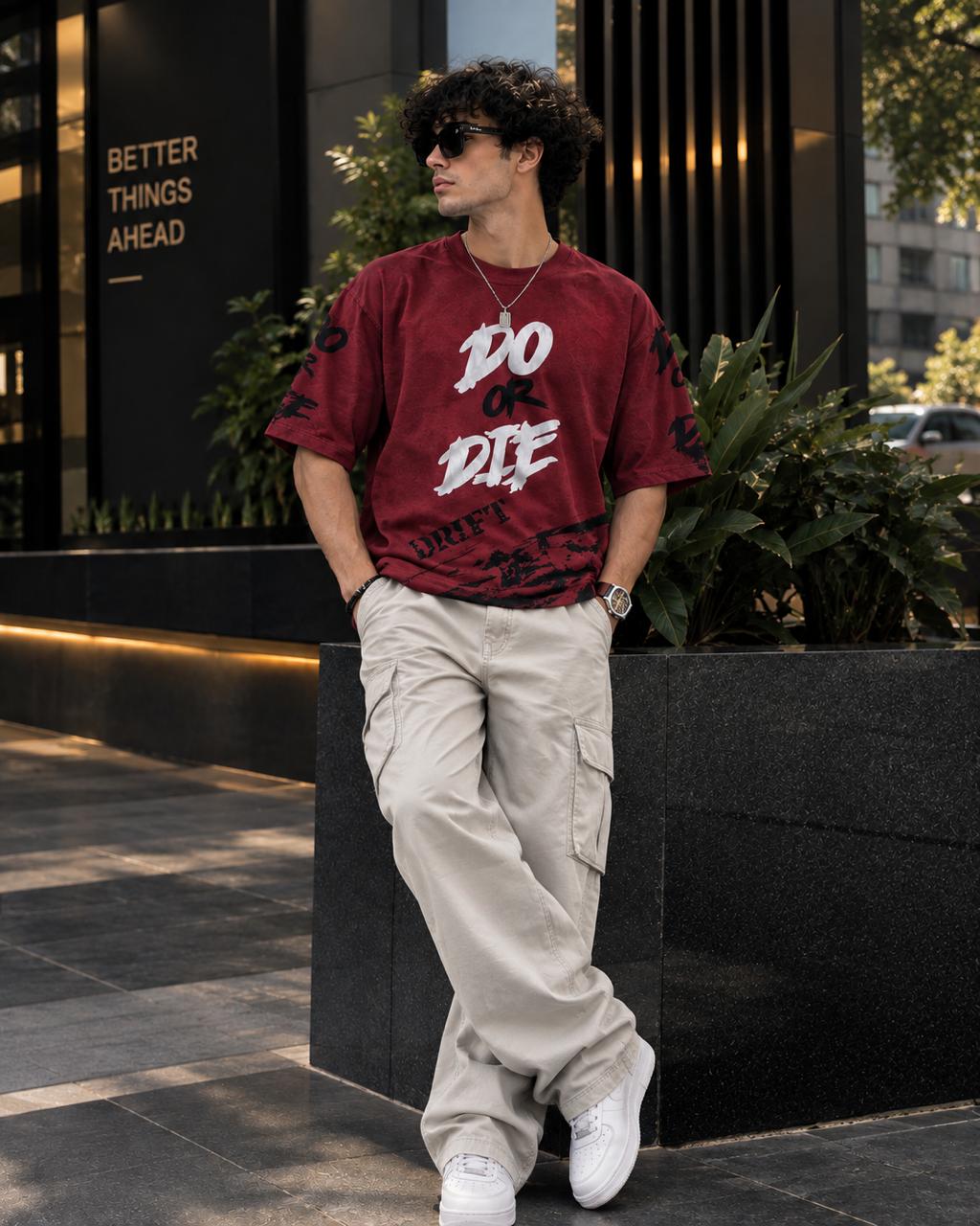 A man wearing a maroon graphic t-shirt with 'DO OR DIE' printed in white, paired with beige cargo pants and white sneakers, leaning against a black stone planter in an outdoor urban setting.