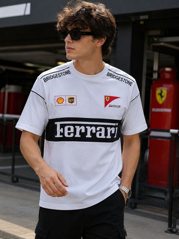 A man wearing a white Ferrari t-shirt with black and red sponsor logos, including Bridgestone, Shell, and UPS, set against a dark garage background with a red Ferrari fire station visible.