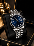 A stainless steel wristwatch with a deep blue dial and date window, resting on a black leather cushion inside a luxury watch box with gold trim.